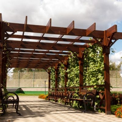 Wooden pergolas from natural wood with comfortable benches in a modern country village on a sunny summer day. The paved path is laid past flower beds, ornamental shrubs and trees.